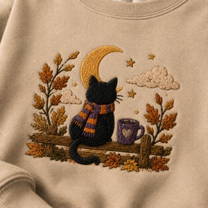 Peut inclure: Un sweat-shirt beige avec une broderie représentant un chat noir portant une écharpe rayée violette et orange, assis sur une clôture en bois. Le motif inclut un croissant de lune jaune, des étoiles, des nuages, une tasse violette et des feuilles.