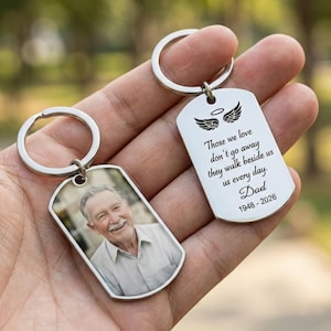 May include: Two silver dog tag keychains. One features a photo of a smiling man. The other has angel wings and the text "Those we love don't go away they walk beside us us every day. Dad 1948-2026."