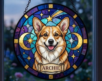 May include: A round stained glass window featuring a smiling corgi dog with brown and white fur. The design includes a blue and purple background with gold stars, moons, and a banner that reads "ARCHIE". The window is suspended by a black chain.