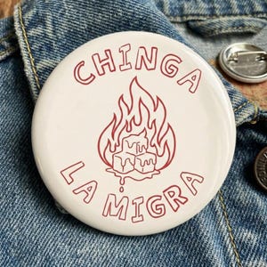 May include: A white button with red text and a graphic of flames and melting ice cubes. The text reads "CHINGA LA MIGRA" in a bold, outlined font. The button is set against a denim background.