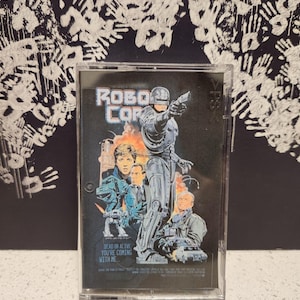 May include: A clear plastic case with a black and white cassette tape inside. The cassette tape has a picture of a cyborg police officer from the movie RoboCop. The text on the cassette tape says "RoboCop" and "Dead or Alive, You're Coming With Me."