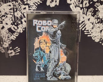 May include: A clear plastic case with a black and white cassette tape inside. The cassette tape has a picture of a cyborg police officer from the movie RoboCop. The text on the cassette tape says "RoboCop" and "Dead or Alive, You're Coming With Me."
