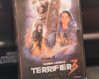 May include: A VHS tape case for the horror movie "Terrifier 3" featuring a clown with a chainsaw and a woman in the background. The text on the case reads "Terror under the Christmas moonlight" and "Damien Leone's Terrifier 3". The release date is listed as "10.11.24".
