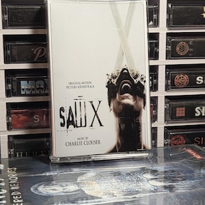 May include: A Saw X original motion picture soundtrack cassette tape featuring a white cover with black text and a graphic of a person with metal devices over their eyes. The cassette is positioned in front of a white storage rack filled with other tapes.