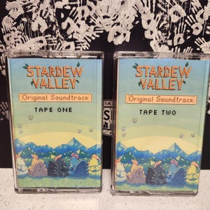 May include: Two clear plastic cases with Stardew Valley Original Soundtrack printed on the front. The left case says "Tape One" and the right case says "Tape Two". Both cases have a colorful image of chickens in a field with mountains in the background.