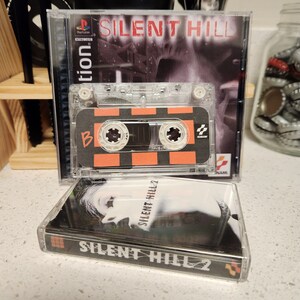 May include: Two video game cases for the Playstation console. The top case is for the game Silent Hill and features a black and white image of a woman's face. The bottom case is for the game Silent Hill 2 and features a black and white image of a woman's face. Both cases have a black background with white text.