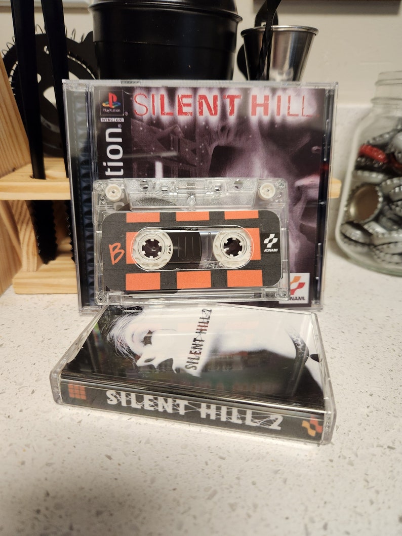 May include: Two video game cases for the Playstation console. The top case is for the game Silent Hill and features a black and white image of a woman's face. The bottom case is for the game Silent Hill 2 and features a black and white image of a woman's face. Both cases have a black background with white text.