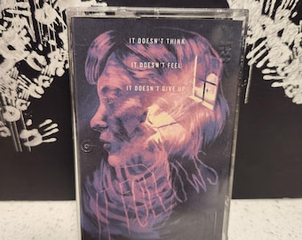 May include: A cassette tape with a black background and a colorful image of a person's face. The text on the tape reads "It doesn't think, It doesn't feel, It doesn't give up".
