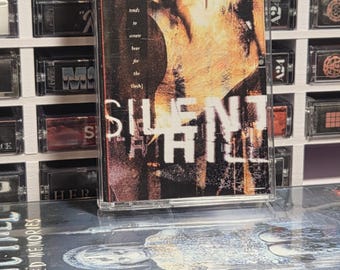 Op de afbeelding: Een Silent Hill-cassettebandje met een donkere, getextureerde hoes met de tekst "the fear of the blood tends to create fear for the flesh" en "SILENT HILL" in het wit, geplaatst voor een Silent Hill Shattered Memories-gamehoes.
