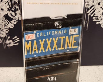 May include: A cassette tape with a blue and yellow California license plate that reads "OCT MAXXXINE CA 85" and the text "A24" on the bottom.