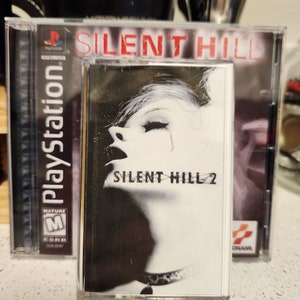 May include: A PlayStation game case for Silent Hill 2. The case is black with a white image of a woman's face and the text "Silent Hill 2". The case is next to a PlayStation game case for Silent Hill. The case is black with a red title "Silent Hill".