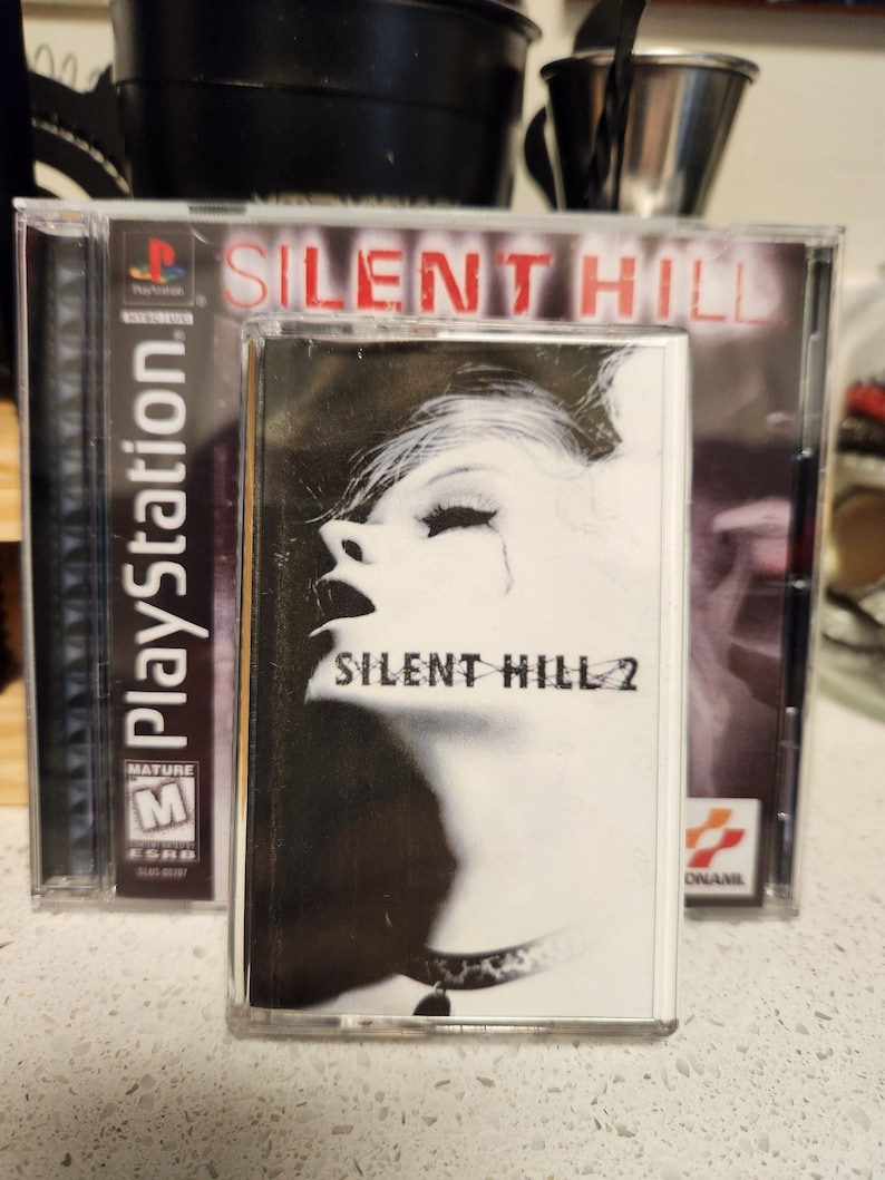 May include: A PlayStation game case for Silent Hill 2. The case is black with a white image of a woman's face and the text "Silent Hill 2". The case is next to a PlayStation game case for Silent Hill. The case is black with a red title "Silent Hill".