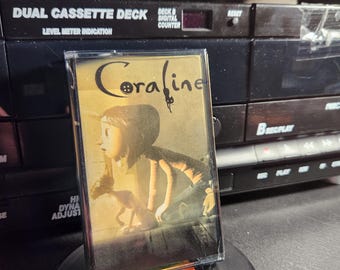 May include: A cassette tape with a yellow and white label featuring the title "Coraline" in a stylized font. The image on the label is a cartoon of a girl with black hair and a red dress standing next to a cat.