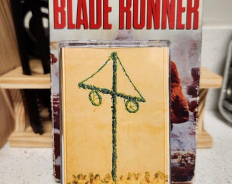 May include: A cassette tape with a yellow and green image of a maypole with people dancing around it. The text "HARRISON FORD IS BLADE RUNNER" is on the cassette case.