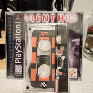 May include: A clear plastic case with a black and orange cassette tape inside. The cassette tape has the words "Silent Hill" printed on it. The case is labeled "PlayStation" and has a "Mature" rating.