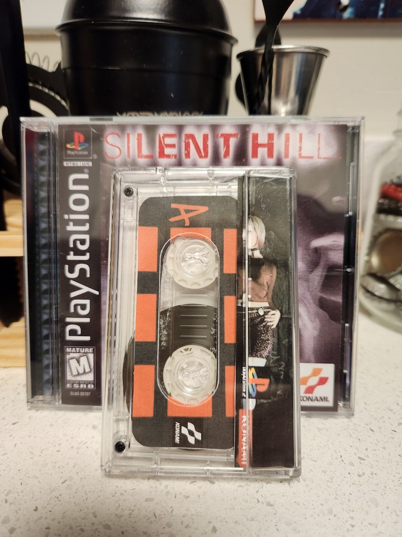 May include: A clear plastic case with a black and orange cassette tape inside. The cassette tape has the words "Silent Hill" printed on it. The case is labeled "PlayStation" and has a "Mature" rating.