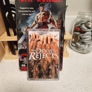 May include: A VHS tape of the movie "The Devil's Rejects" with a red, white, and black color scheme. The tape is in a clear plastic case. The movie is a horror film directed by Rob Zombie. The text on the tape reads "The Devil's Rejects".