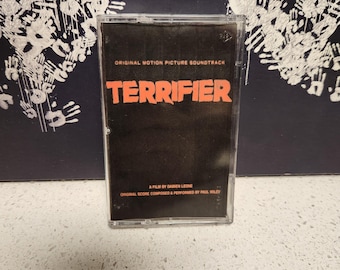 May include: A black cassette tape with the text "ORIGINAL MOTION PICTURE SOUNDTRACK" and "TERRIFIER" in orange. The text "A FILM BY Damien Leone" and "ORIGINAL SCORE COMPOSED & PERFORMED BY PAUL HILEY" is printed in white on the bottom of the cassette.