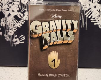 May include: A cassette tape featuring the "Gravity Falls" soundtrack. The cover is brown with the show's logo and a hand with the number 1. The text "Original Score from the Disney Channel Series" and "Music by BRAD BREECK" are also visible.