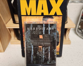 May include: A VHS cassette tape with a black and white image of a wooden cabin in the woods. The text "HEREDITARY" is printed on the cassette tape. The cassette tape is sitting on a table next to a VHS tape case with the text "MEL GIBSON MAD MAX" printed on it.