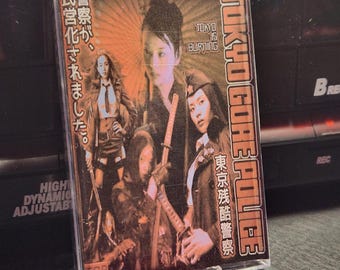 May include: A cassette tape with the title "TOKYO GORE POLICE" in white and red lettering. The cover art features images of women with swords and Japanese text. The tape is in a clear plastic case, resting on a vinyl record.