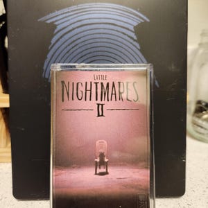 May include: A clear plastic case with a pink and white video game cartridge inside. The cartridge has the text "LITTLE NIGHTMARES II" on the front. The case is sitting on a black surface.
