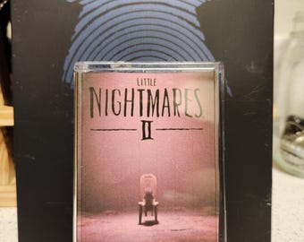May include: A clear plastic case with a pink and white video game cartridge inside. The cartridge has the text "LITTLE NIGHTMARES II" on the front. The case is sitting on a black surface.