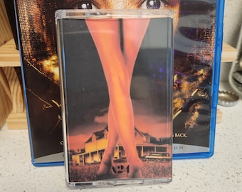 May include: A VHS tape with a black background and a woman's legs in the foreground. The woman's legs are crossed and she is wearing a red dress. The background is a blurry image of a house. The text "Silent Hill" is visible on the VHS tape.
