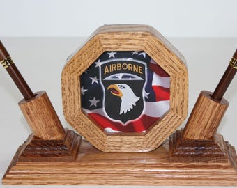 Könnte beinhalten: Schreibtisch-Organizer aus Holz mit zwei Stiften. Der Organizer hat einen achteckigen Rahmen in der Mitte, der eine amerikanische Flagge und das "AIRBORNE"-Emblem zeigt. Die Stifte sind braun mit goldenen Akzenten.