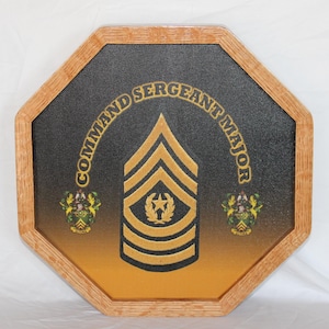 Puede incluir: Placa de madera octogonal con un fondo degradado negro y dorado. La placa presenta las palabras "COMMAND SERGEANT MAJOR" en letras doradas, una insignia dorada y dos escudos. El marco es de madera marrón claro.
