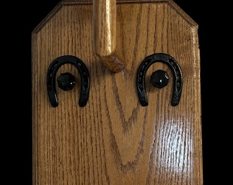 Puede incluir: Perchero de madera para sombreros con dos herraduras negras para colgar sombreros. El perchero está hecho de madera oscura con veta natural.