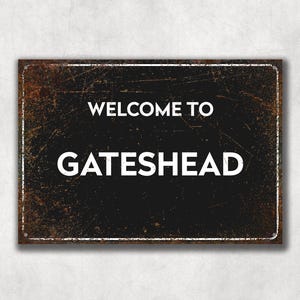 May include: A black metal sign with a rusty texture. The sign reads "Welcome to Gateshead" in white text.