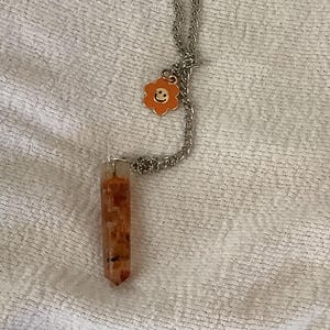 May include: A silver chain necklace with a small orange flower charm and a long, orange, crystal point pendant.