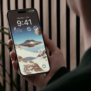 May include: A smartphone displays a futuristic wallpaper featuring a silver flying saucer over a snowy mountain range, with the date and time visible at the top. The phone is held in a person's hand.