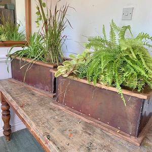 May include: Two rectangular metal planters with wooden edging, filled with green plants, sit on a distressed wooden table. The planters have a reddish-brown colour and are riveted. The table has a weathered finish with paint splatters, typical of a workshop.