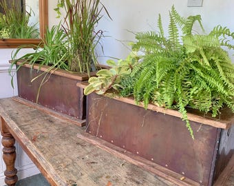 May include: Two rectangular metal planters with wooden trim, filled with various green plants, sit on a weathered wooden table. The planters have a reddish-brown hue and are riveted along the sides. The table has a distressed finish with paint splatters.