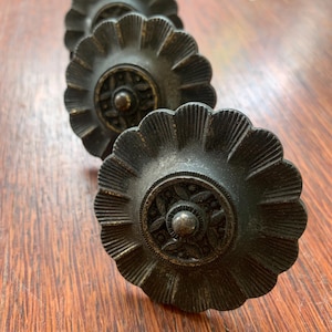 May include: Three antique bronze cabinet knobs are displayed. Each knob has a fluted, flower-shaped design with a detailed centre. The knobs are arranged diagonally on a wooden surface, highlighting their intricate details and aged patina.