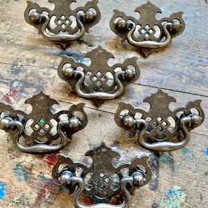 May include: Five antique bronze drawer pulls. Each pull has a decorative backplate with an ornate design and a curved handle. The pulls are arranged on a weathered wooden surface with paint splatters.