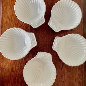 May include: Five white ceramic shell-shaped dishes. Each dish features a ribbed design and a floral accent at the base. The dishes are arranged on a dark wooden surface, highlighting their elegant design.