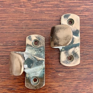 May include: Two antique brass hooks with a rounded edge on one side. The hooks have a worn, aged patina and are mounted on a wooden surface.