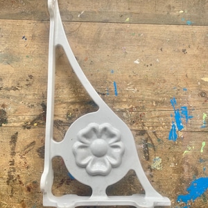 May include: A white, cast iron decorative bracket with a floral design. The bracket is triangular with a rounded floral medallion in the centre. The bracket is designed to be mounted on a wall or other surface.