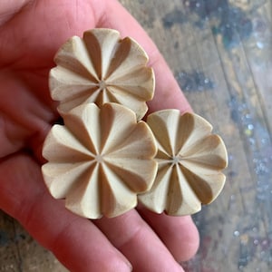 May include: Three small, light-coloured, flower-shaped wooden decorative objects. Each has a central starburst design with radiating petals. The objects are held in a hand, showcasing their size and detail.