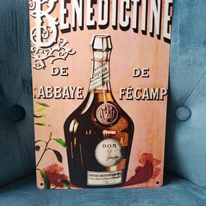May include: Vintage metal sign featuring a bottle of Benedictine liqueur. The sign has a peach-colored background with the brand name in white and the bottle in the center. The bottle is brown with a white label and a detailed design.
