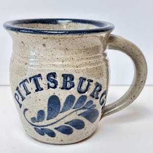 May include: A white ceramic mug with a blue glaze and a blue leaf design. The mug says "PITTSBURG" in blue lettering.