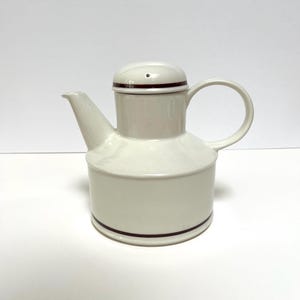 May include: White ceramic teapot with a brown stripe around the top and bottom. The teapot has a lid with a small hole in the top.