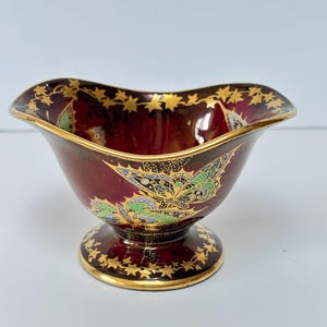 May include: A burgundy pedestal bowl with a scalloped rim. It has gold leaf detailing on the rim and base, and a detailed floral and leaf pattern in green, blue, and gold. The bowl is likely ceramic or porcelain.