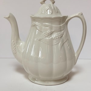 Może przedstawiać: Biały ceramiczny czajnik z pokrywką i uchwytem. Czajnik jest ozdobiony wzorem pszenicy.