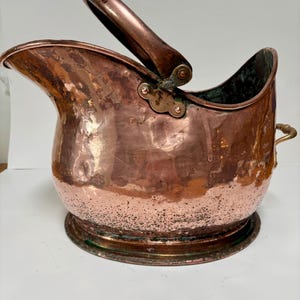 May include: A copper coal scuttle with a hinged handle and brass accents. The scuttle has a hammered finish and a rounded base.