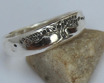 May include: A silver ring with a tree design engraved on the band. The tree has a thick trunk and branches with leaves. The ring is on a small rock.