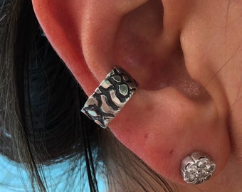 May include: A silver ear cuff with a textured design and a small silver stud earring with a clear stone.